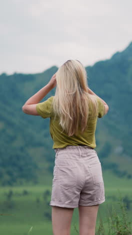 female tourist adjusts shorts and shakes loose blonde hair breathing fresh air against mountains with pine forest in nasty weather backside view slow motion