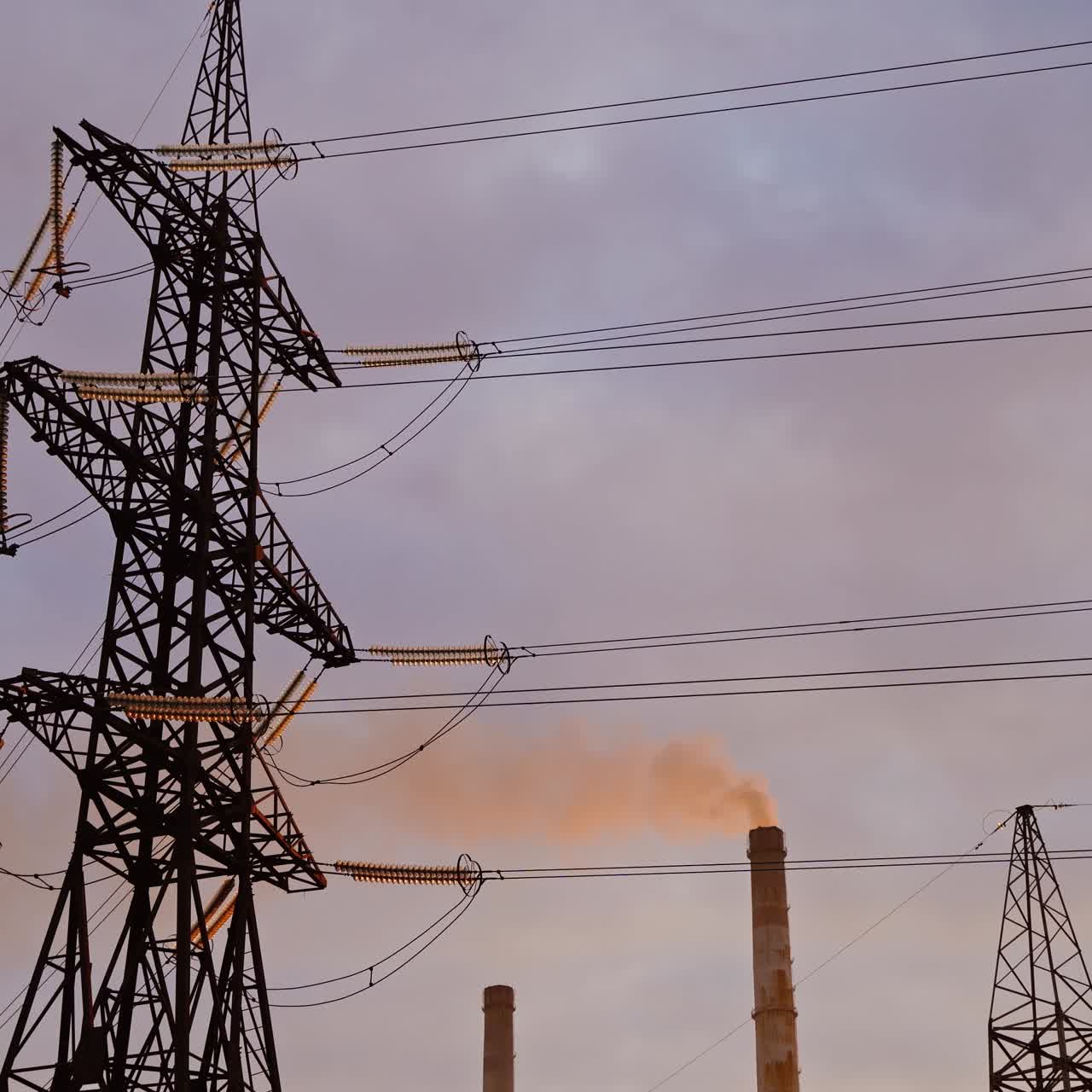 Electricity pylon on industrial chimney background. High-voltage tower and pipe of power plant with smoke on evening sky. Harmful smoke polluting the environment.
