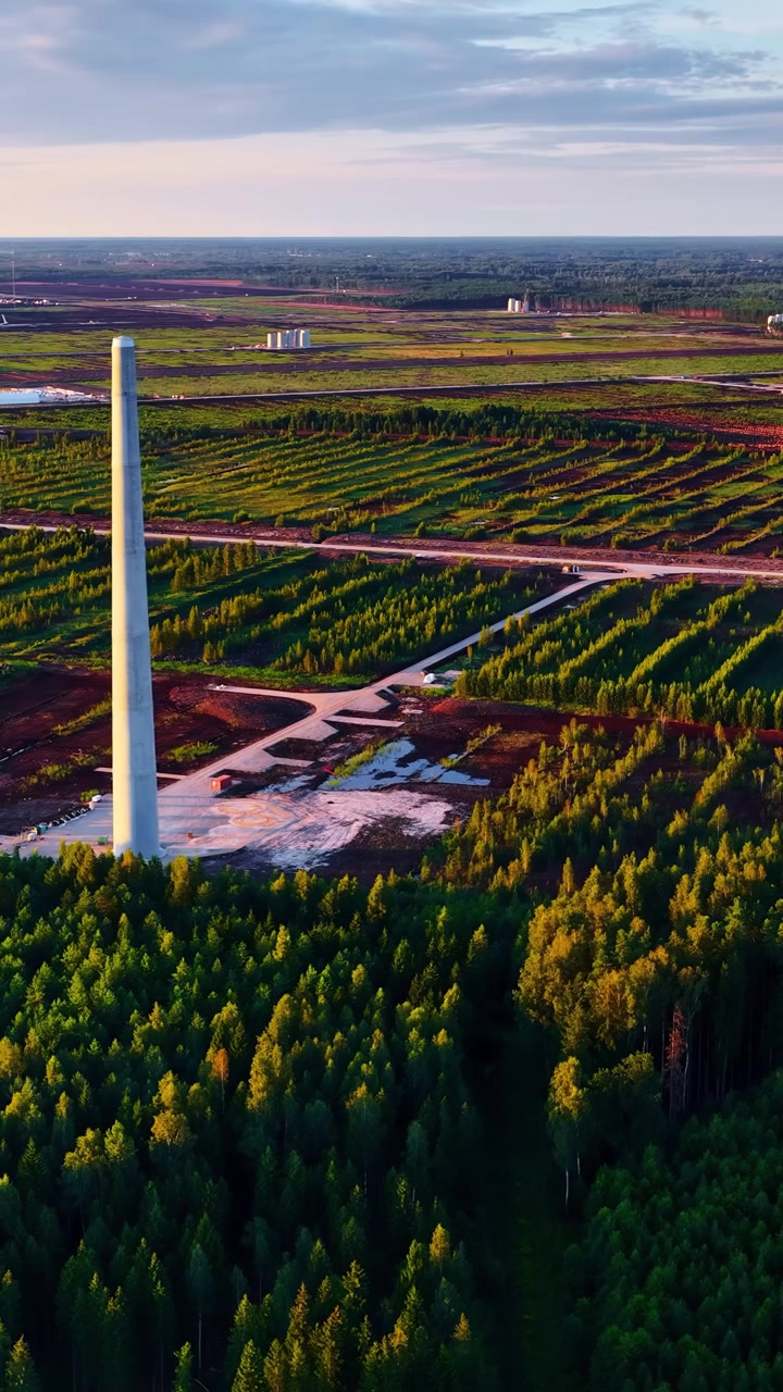 A tall chimney stands amidst a patchwork of fields and dense forest under a setting sun. Ideal for environmental documentaries or industrial themes.