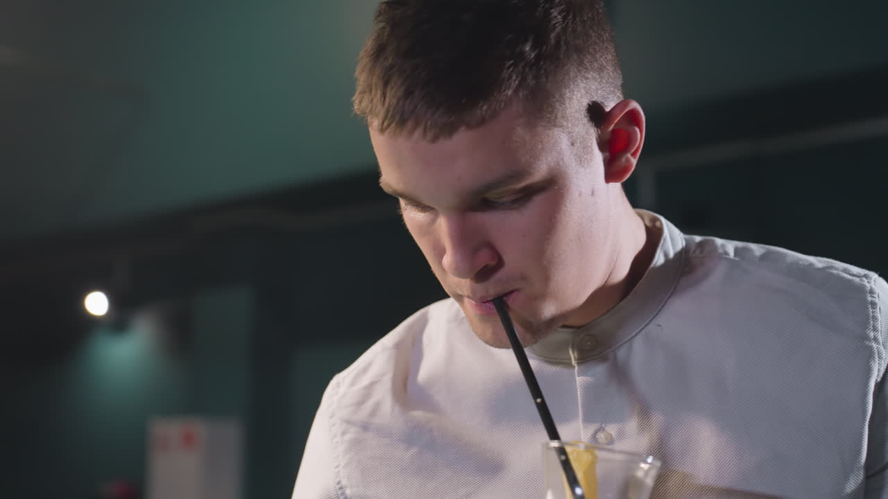 primer plano de un joven con camisa blanca bebiendo una bebida de limón con paja negra en una habitación débilmente iluminada. la iluminación ambiental proyecta una atmósfera de mal humor, enfatizando la expresión facial y la postura relajada