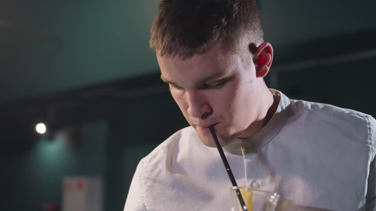 Primo piano di un giovane uomo in camicia bianca che sorseggia una bevanda al limone con una cannuccia nera in una stanza scarsamente illuminata. L'illuminazione ambientale crea un'atmosfera suggestiva, enfatizzando l'espressione del viso e la postura rilassata.