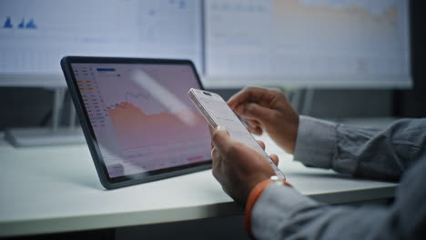 african american financial analyst checking real-time stocks, exchange market charts on smartphone and digital tablet using online trading app. businessman working in investment bank office. close up.
