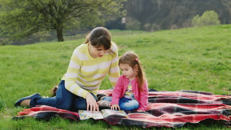 Eine-Junge-Frau-Sitzt-Auf-Dem-Grünen-Gras-Und-Bringt-Ihrer-Tochter-Bei,-Ein-Buch-In-Der-Natur-Zu-Lesen
