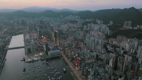 Toma-Panorámica-Aérea-Del-área-De-La-Bahía-De-Kowloon,-Hong-Kong-Después-Del-Atardecer-Con-La-Silueta-De-La-Cordillera-En-El-Fondo