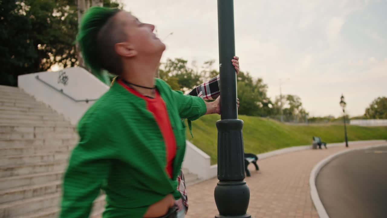 Two lesbian girls with short haircuts of bright colors in pink and green checkered shirts, during their date, spin around a lamppost and continue to walk further holding hands in the park