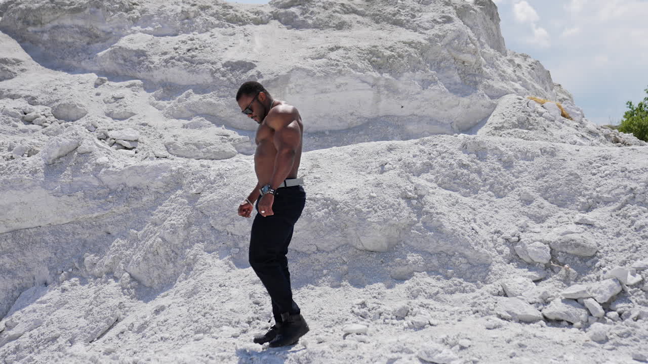 Vista di una telecamera in movimento di un uomo americano allegro a torso nudo che cammina sulla spiaggia sabbiosa. Uomo muscoloso attraente e affascinante con concetto di torso nudo