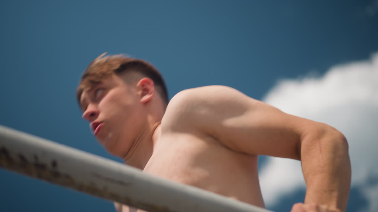 밝은 파란 하늘을 배경으로 야외 철봉에서 턱걸이를 하는 젊은 남성, 클로즈업 카메라 앵글로 힘든 노력과 호흡 포착, 지구력을 밀어붙이는 운동선수 훈련생, 코치와 함께 공원 운동 분위기