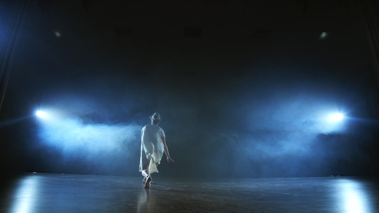 a young girl in a white dress dances contemporary in slow motion on the stage with smoke in spotlights. Camera stands behind her filming in a full shot.