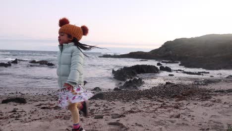 a little girl in a beani dances on a cold coastal beach