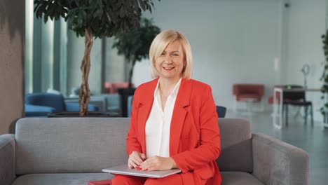 waist-up portrait of senior woman in red suit looking at camera and smiling on office background