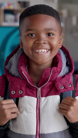 a young african american boy smiles at the camera while wearing a backpack in a school setting.