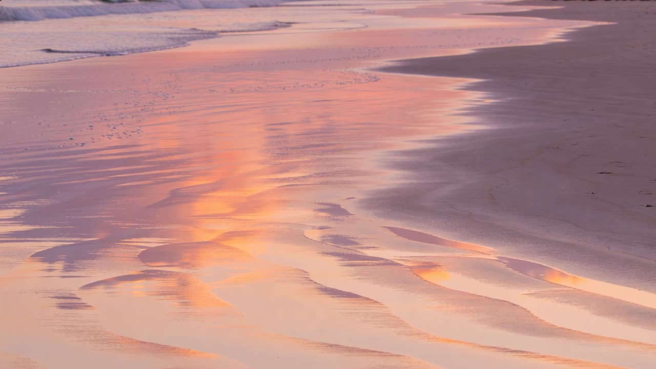 Wet sandy shoreline reflecting warm sky, gentle waves nudging foam and reshaping ripples for travel