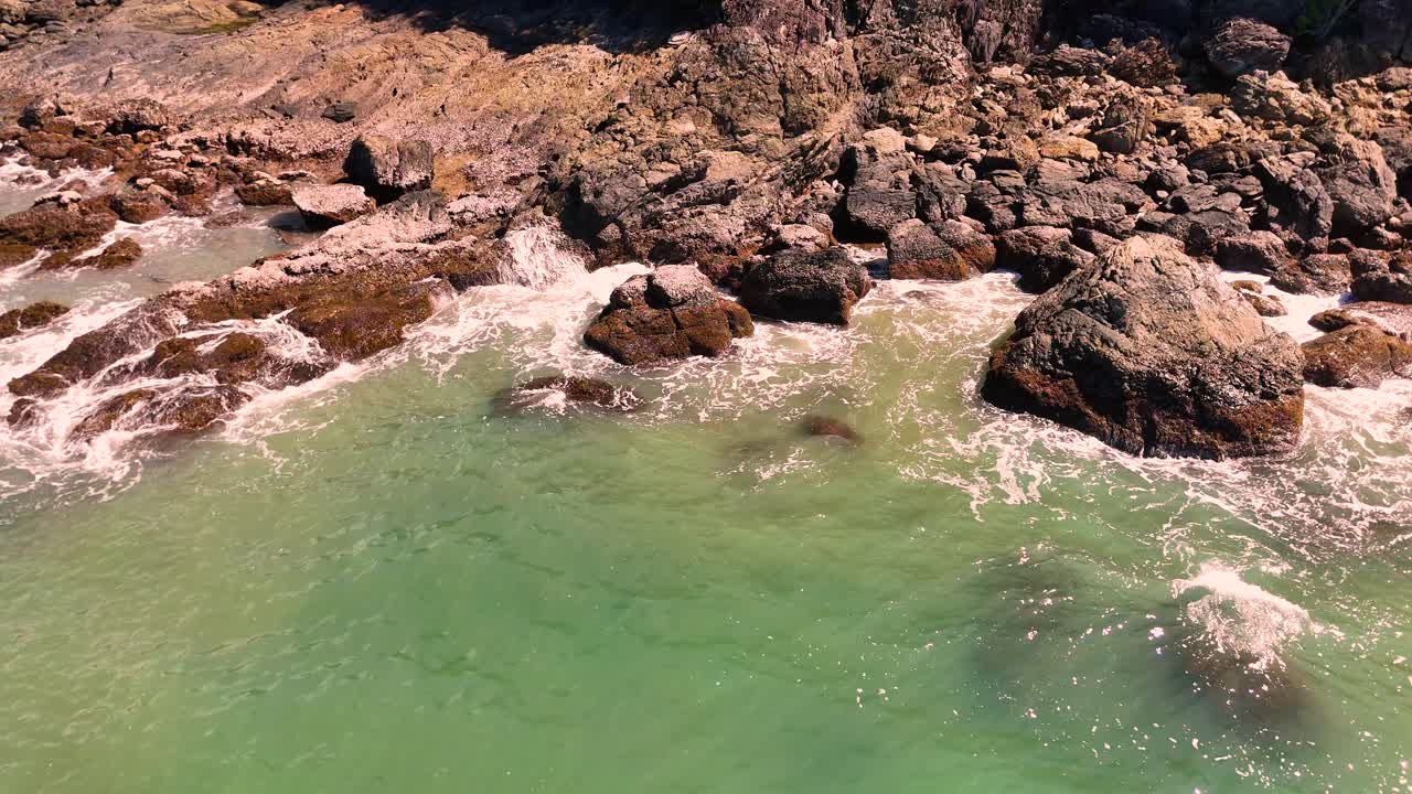 Riprese aeree sorvolano una costa rocciosa con acqua turchese, onde leggere e una luminosa luce solare naturale, catturando un ambiente tropicale tranquillo