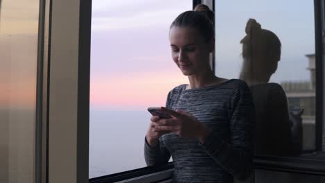 young attractive woman texting a message while standing by the open window with a smile during the sunset by the sea. beautiful sky and sea on the background