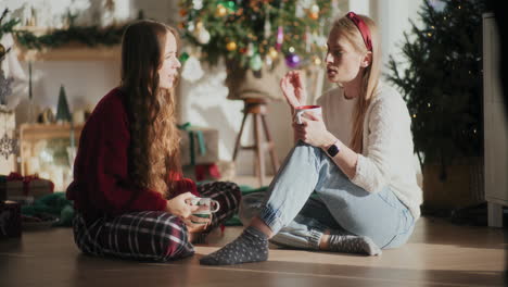 beautiful sisters with coffee cups talking on floor at home during christmas