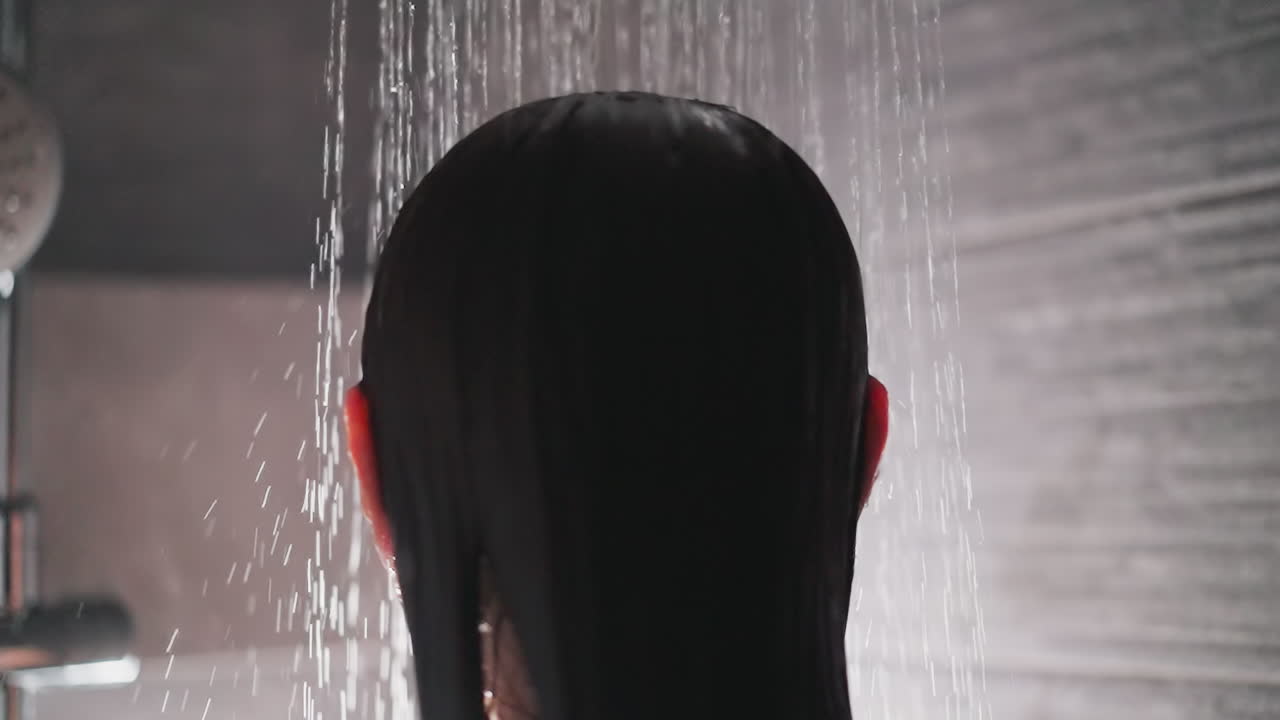 la mujer se da la vuelta con la cabeza bajo el flujo de agua. la dama se mueve con la cabeza con el cabello moreno bajo el chorro de agua en la ducha con el gris de la pared de azulejos
