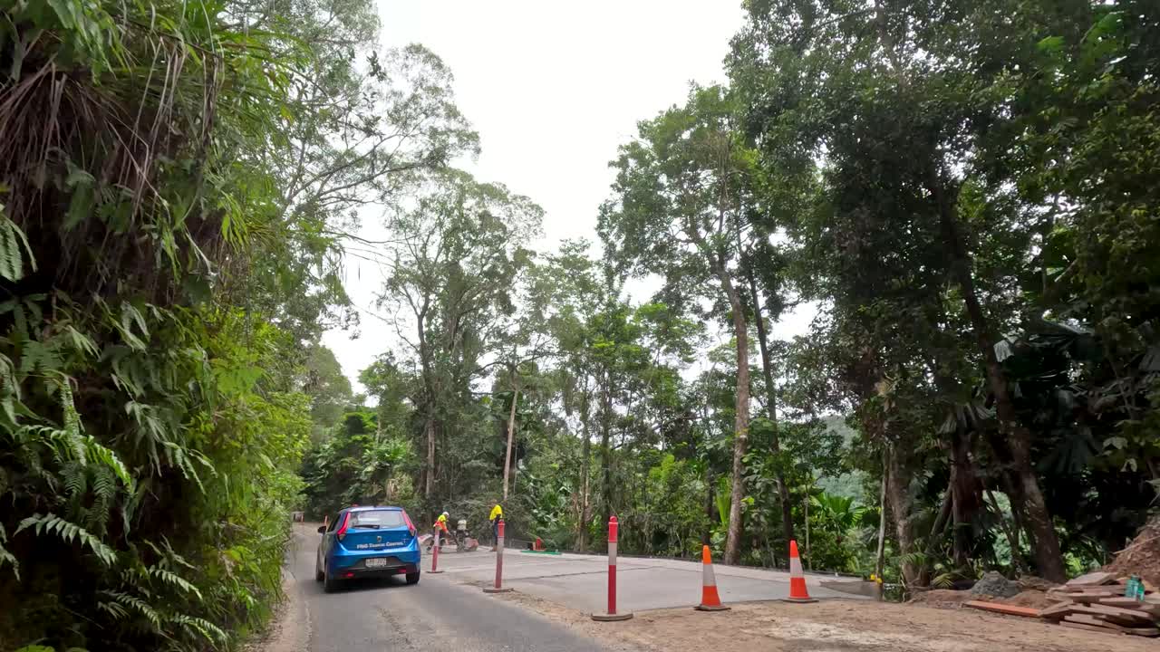 Blue car drives through tropical rainforest roadworks, encountering construction equipment, workers, and traffic cones