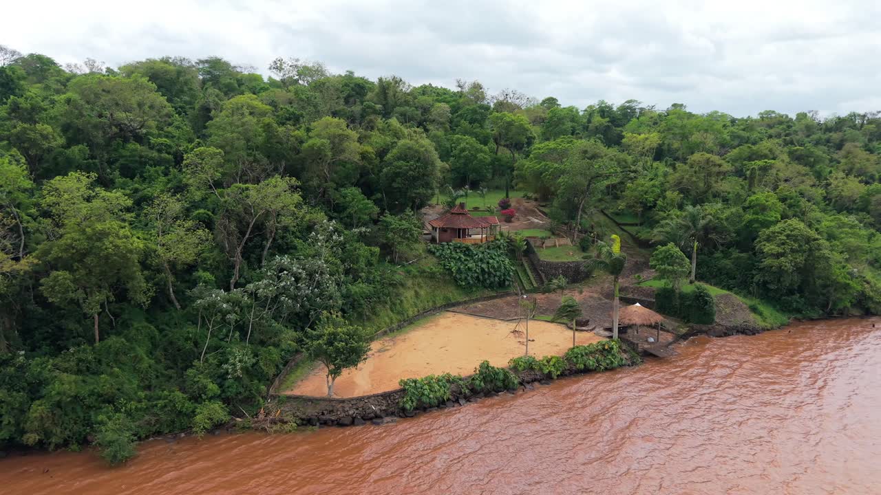 Drone aerial showing a secluded sandy beach and forest compound on the Paraná River, Campichuelo, Paraguay, with lush vegetation and riverbank landscaping, serving as recreational or docking zone