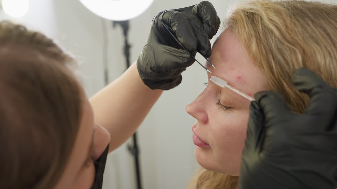 Beautician in black gloves carefully arranges blond client eyebrow using small grooming brush while client keeps eyes closed during beauty session in bright salon, showing attention to detail