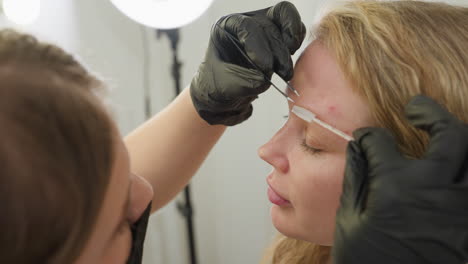 beautician in black gloves carefully arranges blond client eyebrow using small grooming brush while client keeps eyes closed during beauty session in bright salon, showing attention to detail