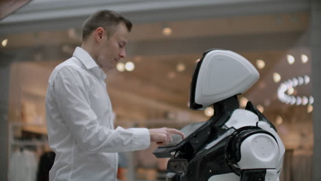 a man in a shirt communicates with a white robot asking questions and pressing the screen with his fingers