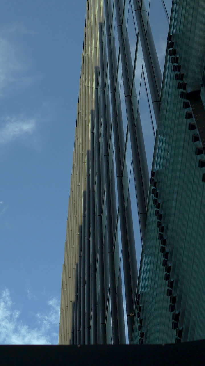 Una llamativa toma vertical en contrapicado de un moderno edificio de oficinas de cristal contra un cielo azul, perfecta para contenido corporativo y arquitectónico.