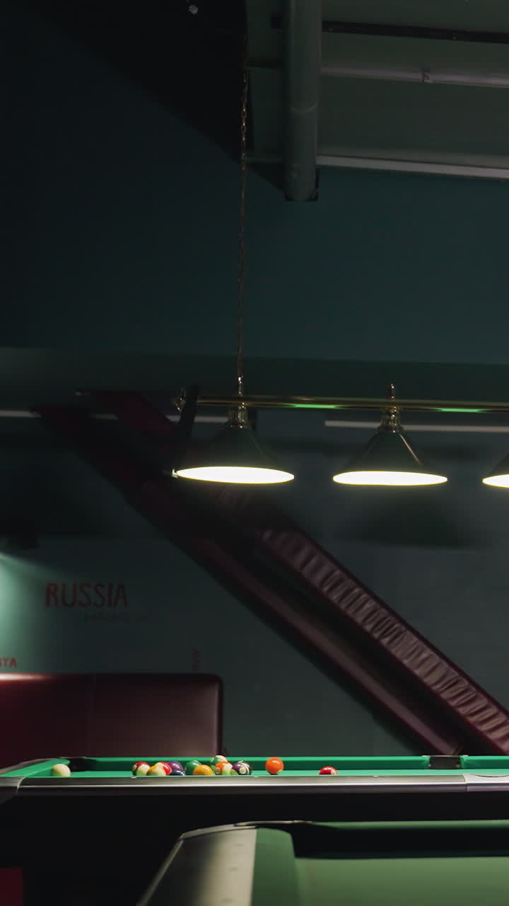 Signore in camicia bianca si china su un tavolo da biliardo verde, mirando a un tiro preciso in una stanza scarsamente illuminata con pareti verde acqua scuro, illuminata da luci calde dall'alto, creando un'atmosfera rilassata ma intensa per il gioco.