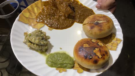 Top-shot-of-a-man-is-seen-in-a-video-holding-a-plate-of-savory-Litti-Chokha-that-has-been-coated-in-ghee-and-is-served-with-roasted-eggplant-bharta-or-baingan-and-a-hot,-spicy-green-chutney