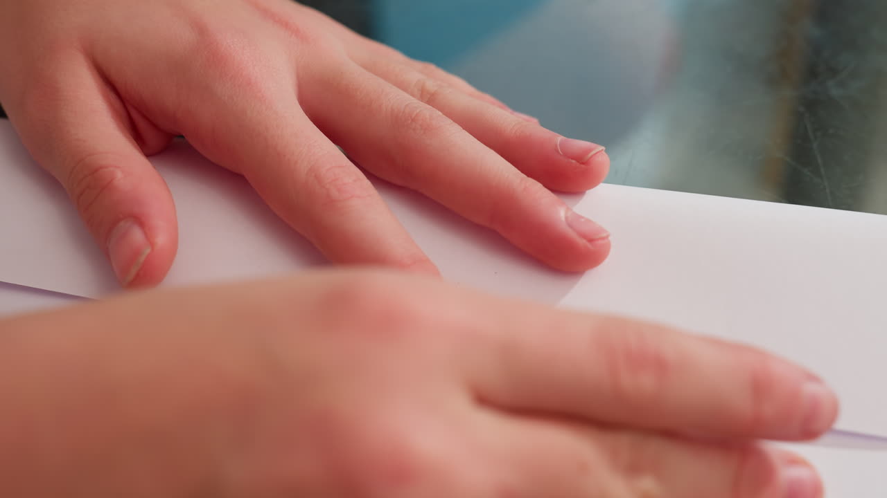 Close-up of hand folding paper carefully, straightening edges on glass table to create paper craft, focusing on precision and neatness