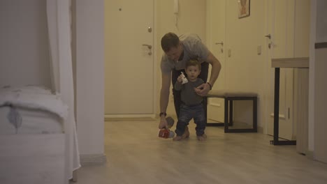 father helping her baby to take his first steps from the corridor to the living room at home