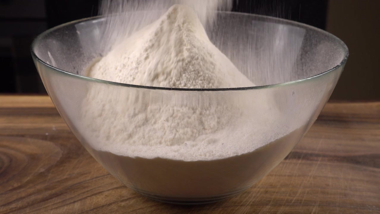 Mengayak tepung menggunakan saringan ke dalam mangkuk kaca di dapur.