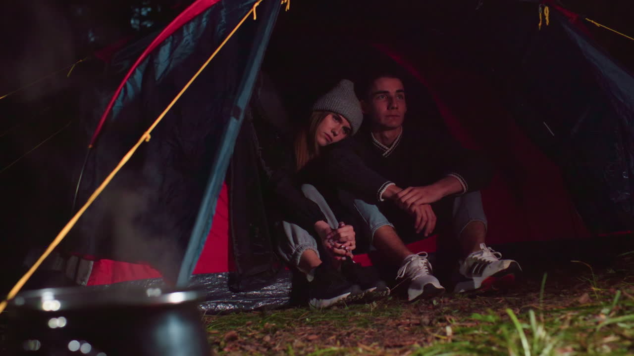 Couples seated close together at entrance of tent during night camping, quietly resting while cooking pot simmers on fire nearby, surrounded by natural forest setting under soft, warm light