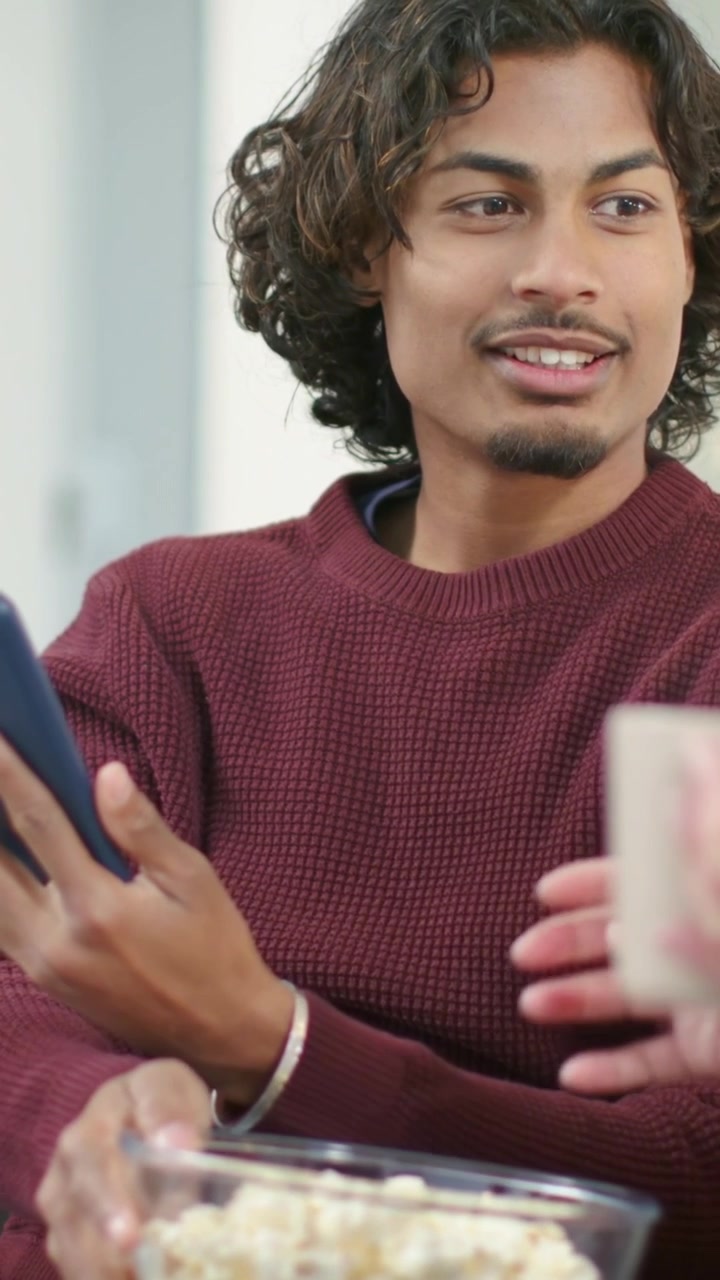 Video verticale: Uomo con maglia bordeaux sul divano che parla, mangia popcorn e controlla il tablet accanto a una tazza floreale.