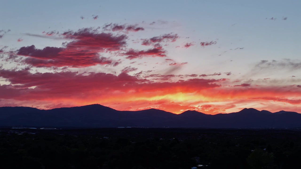 A drone timelapse capturing dramatic orange and red clouds over a dark mountain range after sunset. Vibrant colors illuminate the evening sky as twilight deepens across the rugged landscape.