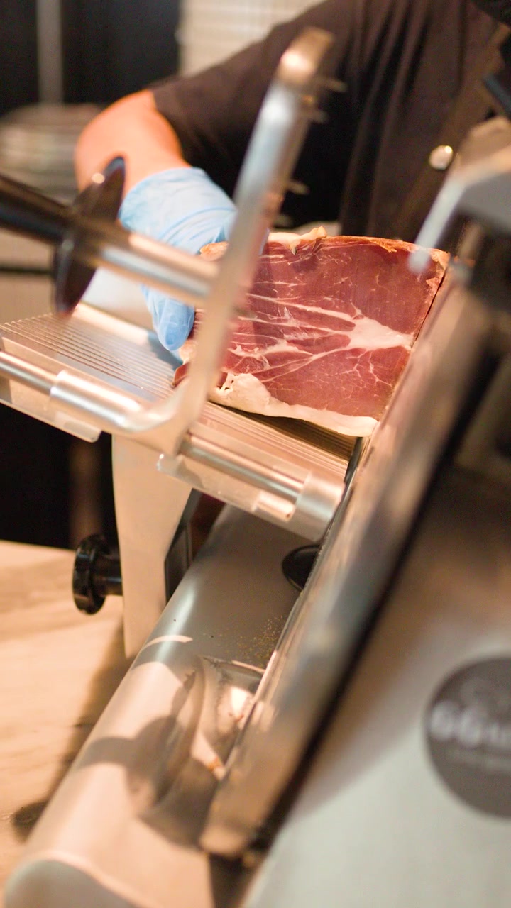Close-up of chef in blue gloves using electric slicer for thin prosciutto meat slices.