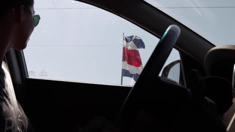 Young-female-model-watching-the-Costa-Rica-flag-gently-waving-in-the-wind,-outside-in-the-street-from-a-car,-slow-motion