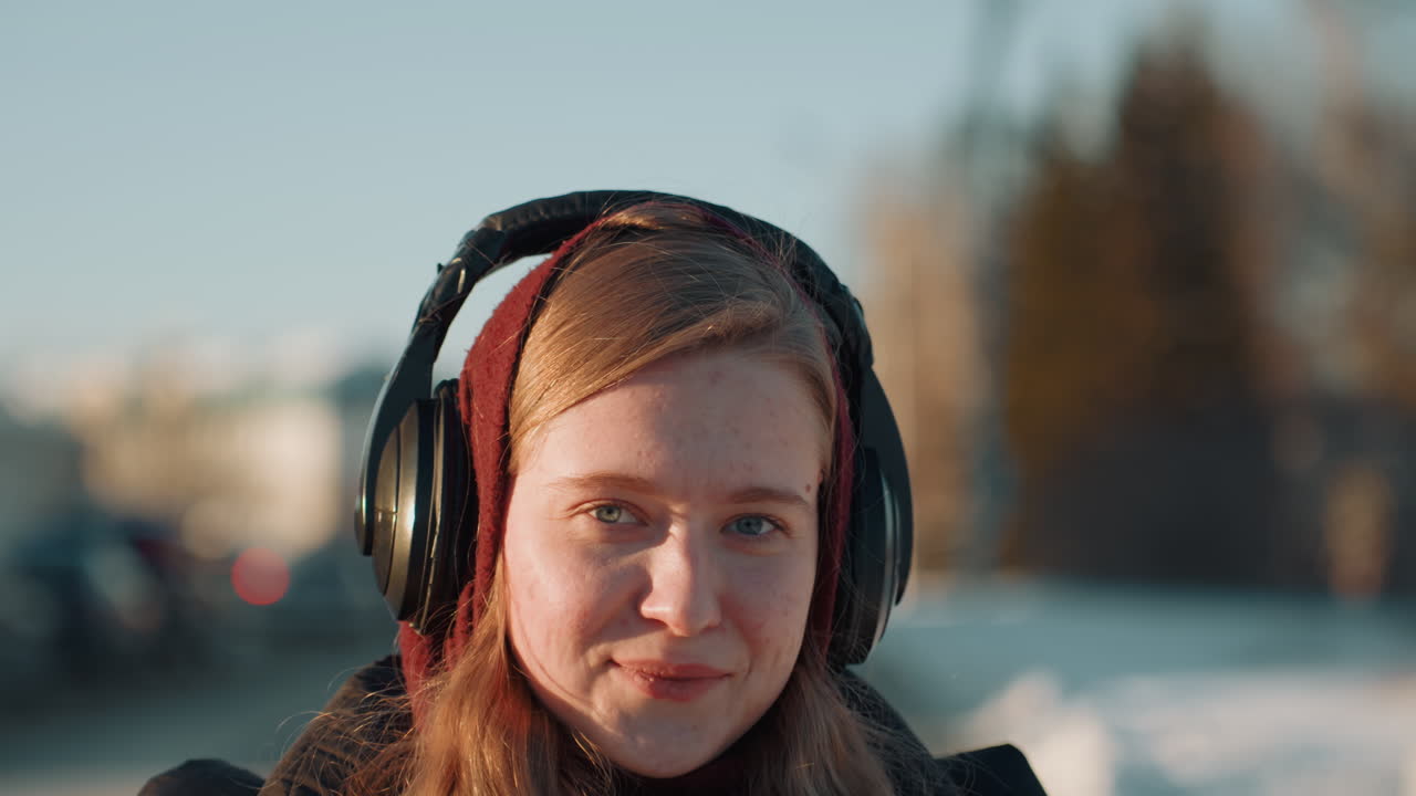 Giovane donna che ascolta musica all'aperto all'ora d'oro, con le cuffie, fiocchi di neve sul cappotto, sguardo verso l'alto e sorriso dolce, luce solare che illumina i capelli, atmosfera invernale pacifica e umore riflessivo.