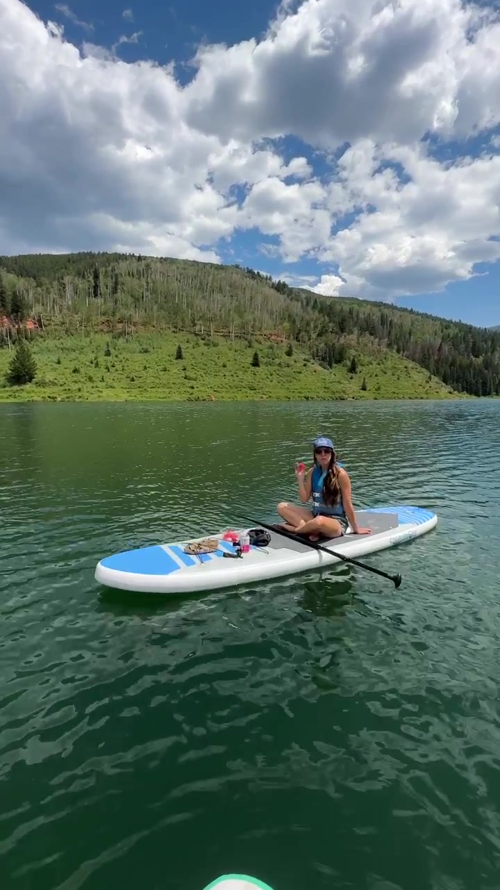 Vista verticale di una giovane donna che mangia e siede su una tavola da paddleboard in una soleggiata giornata estiva, Lago Sylvan, Colorado, USA.