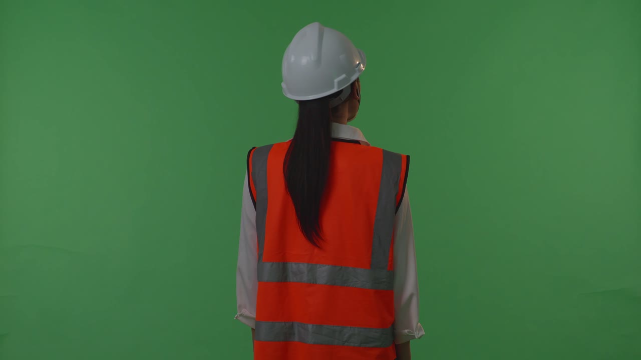 동양의 여성 엔지니어가 안전 헬을 착용하고 초록색 스크린 배경 스튜디오에 서 있는 동안 주변을 둘러보고 있는 뒷면