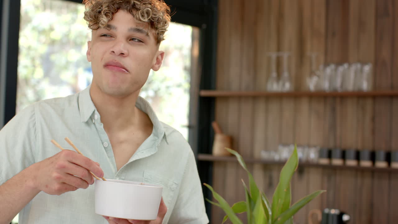 Young man enjoying noodles with chopsticks at home, looking content, copy space