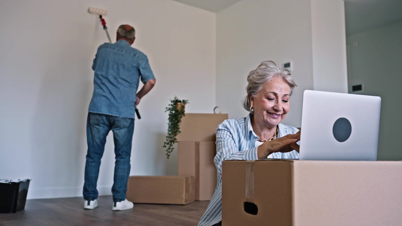 Senior Couple Renovating New Home, Woman Using Laptop While Man Paints Wall