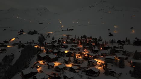 Toma-Aérea-Acercándose-A-La-Ciudad-De-Arosa-En-Una-Montaña-Nevada-Por-La-Noche-En-Suiza