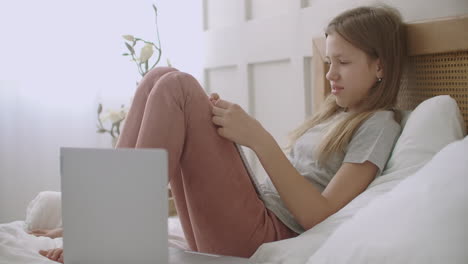 cheerful schoolgirl is talking and smiling sitting on bed with copybook after online learning session e-learning