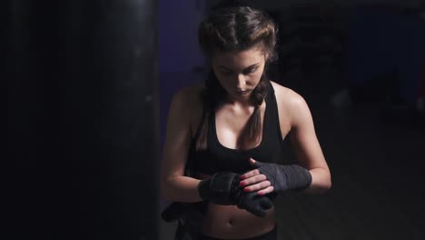 Woman-taking-on-boxing-gloves-on-hands-with-black-boxing-wraps-in-dark-room.-Slow-Motion-shot