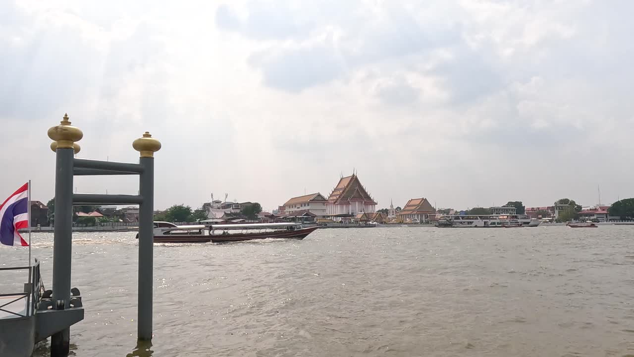 A serene river scene in Bangkok with boats passing by, under a cloudy sky. The camera captures a steady view of the riverbank