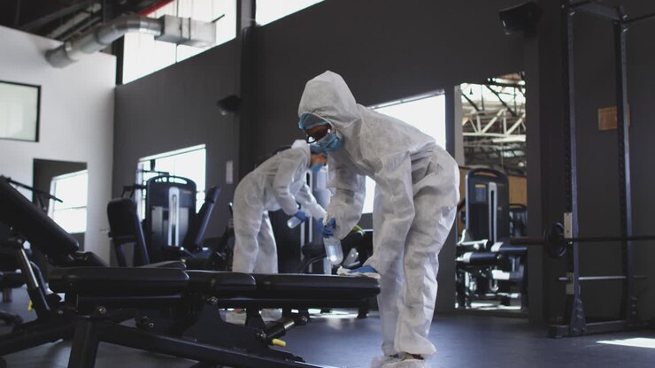 Squadra di operatori sanitari che indossano indumenti protettivi e puliscono una palestra usando uno spruzzatore disinfettante