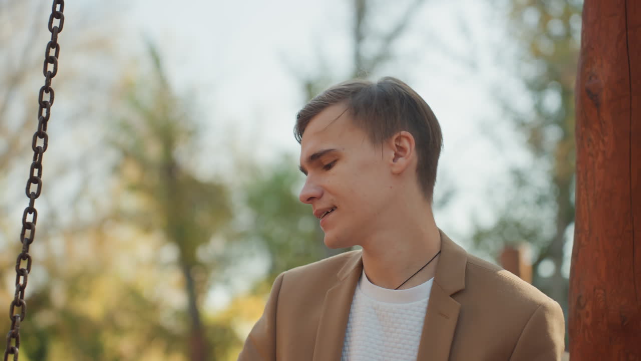 Giovane uomo caucasico sull'altalena, ritratto contemplativo in un parco autunnale che indossa un blazer, sorriso sottile e sguardo pensieroso, espressioni mutevoli suggeriscono memoria, attesa, ricordo di promesse, immaginazione di una famiglia