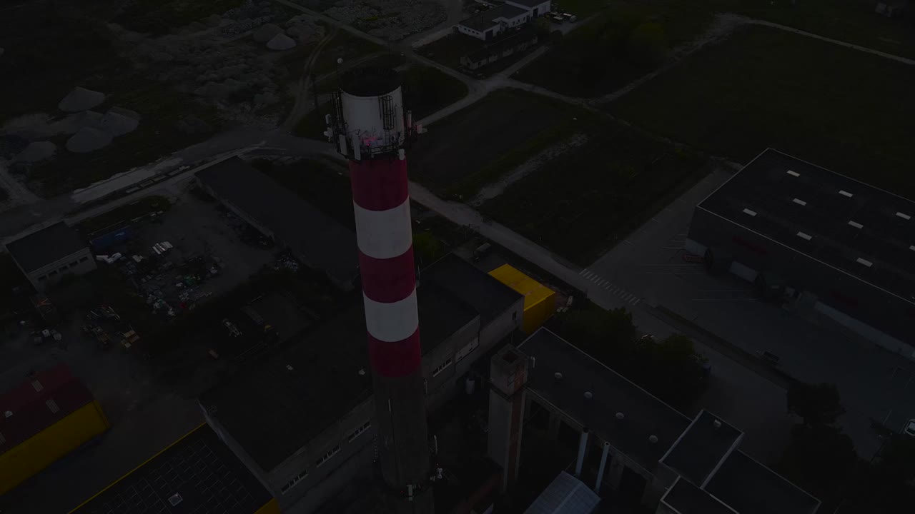 Gorgeous aerial drone revealing footage of a tall factory red and white striped chimney stack on a rural city during orange colored sunset in the summer time with industrial buildings surrounding it.