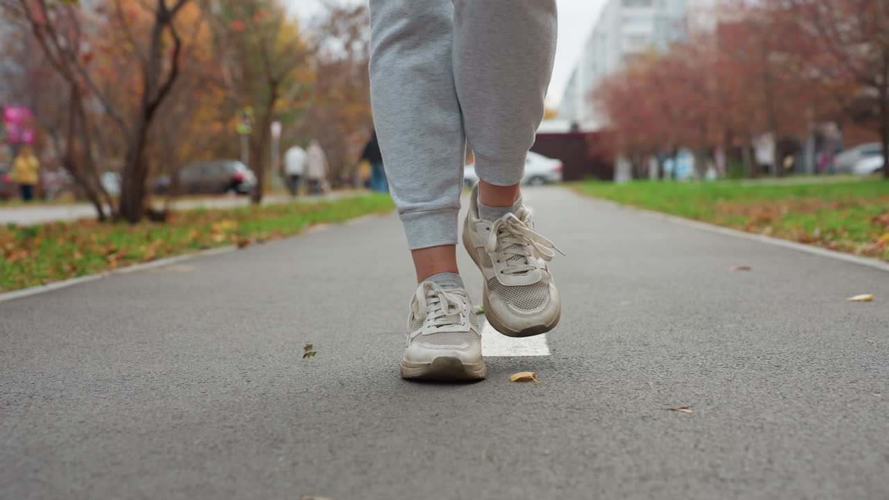 Person in joggers and canvas sneakers walking along white road marking as dry autumn foliage sways with wind, blurred people moving in background, tree-lined path in urban park under cloudy sky