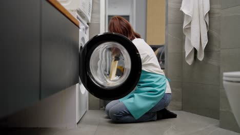 side view of a confident brunette cleaning lady girl in a white t-shirt and blue apron loading things into the washing machine and starting it while cleaning on call